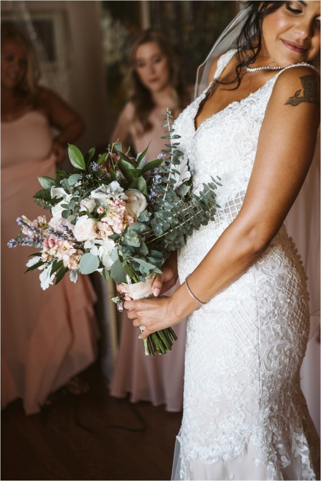 Premier snohomish & seattle wedding photographer | gsquared weddings photography a bride in a lace wedding dress holds a bouquet of pastel flowers and eucalyptus smiling slightly with two bridesmaids in blush dresses standing blurred in the background at this lovely snohomish seattle wedding | serving snohomish seattle                   & beyond