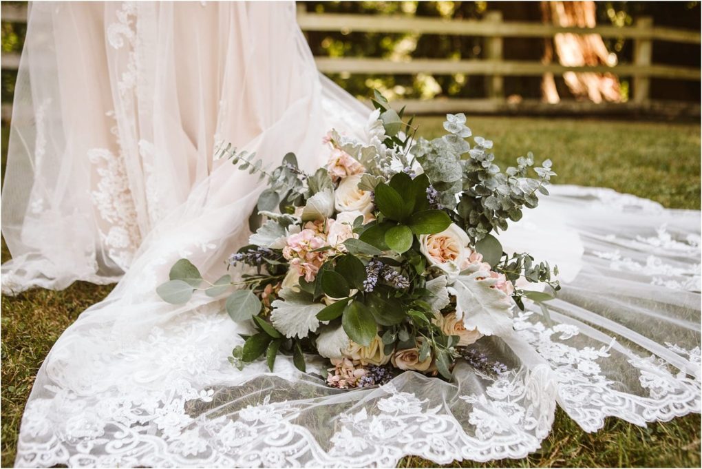 Premier snohomish & seattle wedding photographer | gsquared weddings photography a bridal bouquet of pastel roses greenery and lavender rests on the lace train of a wedding dress in snohomish with grass underfoot and a wooden fence and trees framing this dreamy seattle area wedding scene | serving snohomish seattle                   & beyond