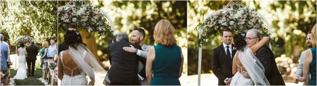 Premier snohomish & seattle wedding photographer | gsquared weddings photography a bride in a white gown and veil stands with her father who hugs and then hands her off to the groom beneath a floral arch during a romantic snohomish outdoor wedding ceremony Guests and officiant look on | serving snohomish seattle                   & beyond