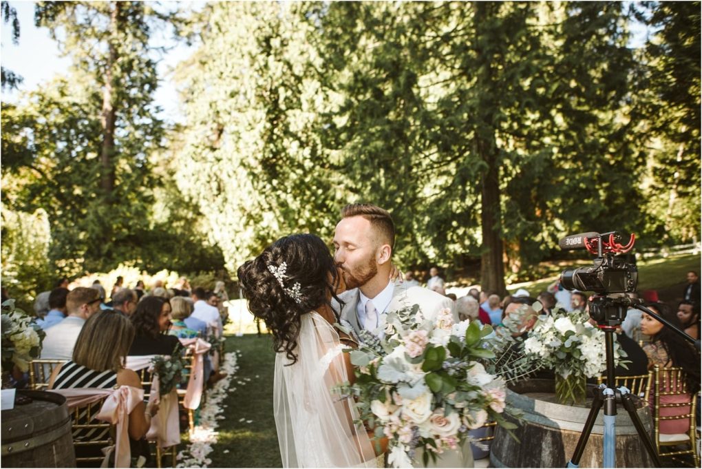 Premier snohomish & seattle wedding photographer | gsquared weddings photography a bride and groom share a kiss outdoors during their snohomish wedding ceremony surrounded by guests on chairs The couple holds a bouquet of flowers with lush greenery and tall trees filling the background | serving snohomish seattle                   & beyond