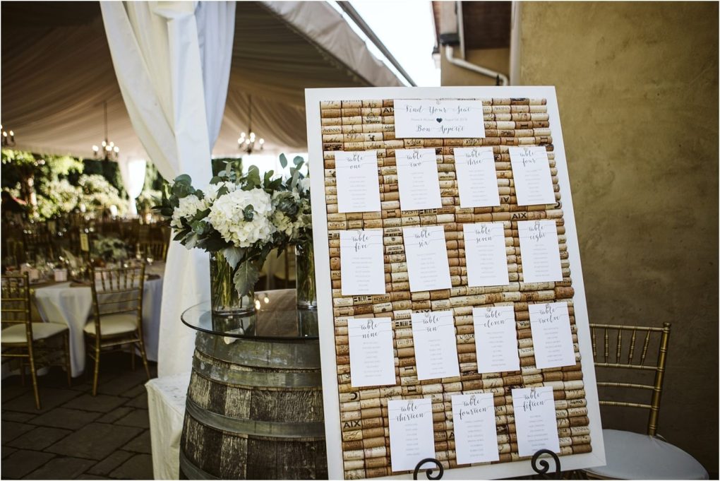Premier snohomish & seattle wedding photographer | gsquared weddings photography a snohomish wedding seating chart made from wine corks and white cards is displayed on an easel next to a wine barrel with a floral arrangement under a tent with set tables and gold chairs | serving snohomish seattle                   & beyond
