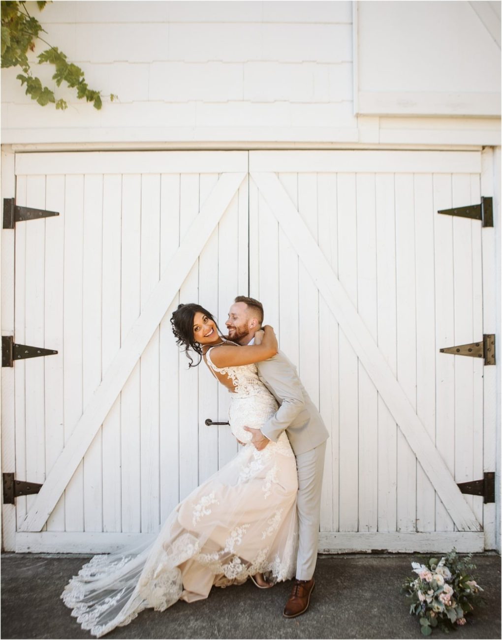 Premier snohomish & seattle wedding photographer | gsquared weddings photography a groom in a light suit lifts and embraces his bride dressed in a lace gown in front of large white barn doors at their snohomish seattle wedding Both are smiling joyfully as a bouquet of flowers rests on the ground nearby | serving snohomish seattle                   & beyond