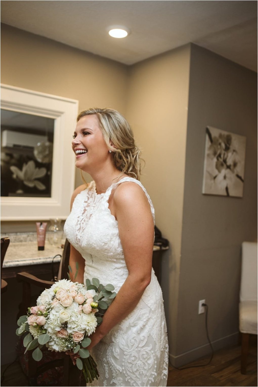 Premier snohomish & seattle wedding photographer | gsquared weddings photography a bride in a white lace wedding dress smiles and holds a bouquet of white and blush flowers in a softly lit snohomish room with beige walls and a mirror capturing the elegance of a seattle wedding engagement | serving snohomish seattle                   & beyond