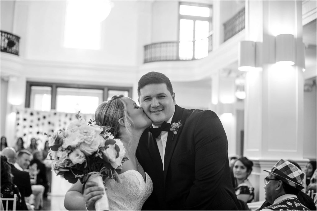 Premier snohomish & seattle wedding photographer | gsquared weddings photography a bride in a strapless wedding dress kisses her smiling groom on the cheek while holding a bouquet They stand together in a bright elegant snohomish venue with guests seated in the background capturing their seattle wedding bliss | serving snohomish seattle                   & beyond
