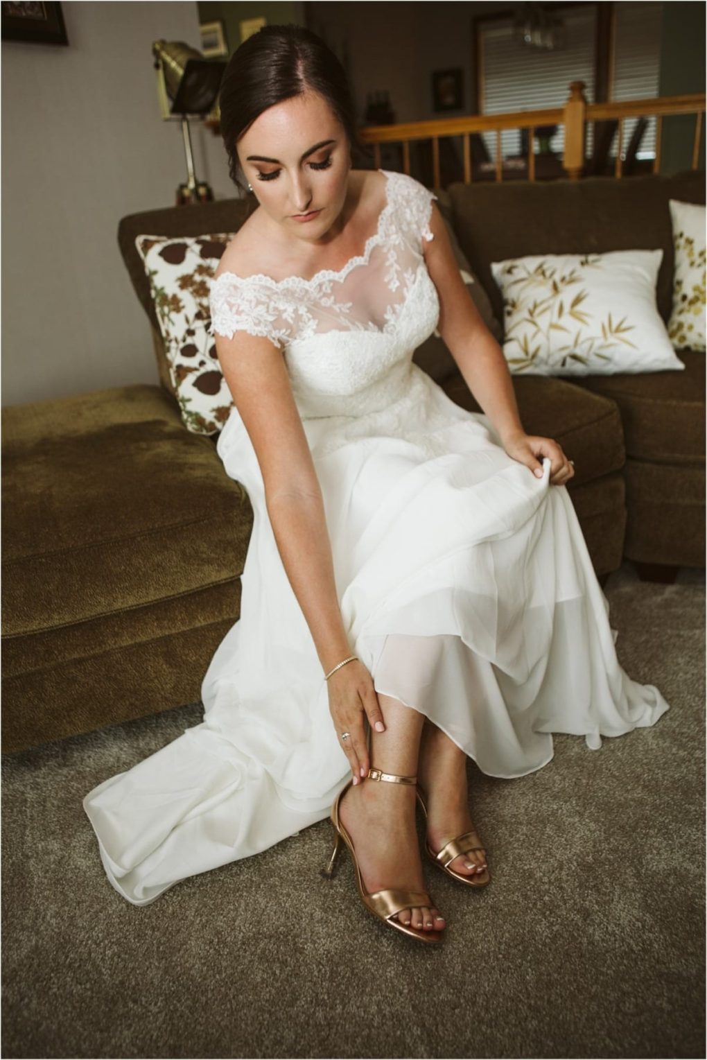 Premier snohomish & seattle wedding photographer | gsquared weddings photography a bride in a white lace wedding dress sits on a couch looking down as she fastens the strap on her gold high heeled sandal This intimate snohomish seattle wedding moment unfolds in a cozy living room with patterned pillows in the background | serving snohomish seattle                   & beyond