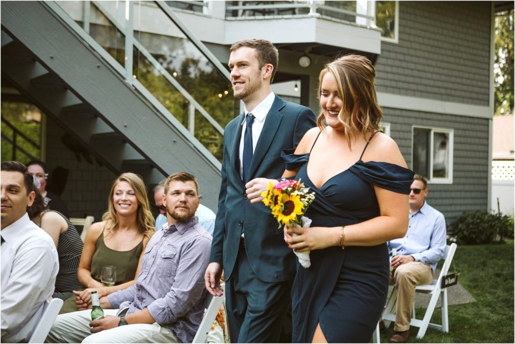 Premier snohomish & seattle wedding photographer | gsquared weddings photography a man in a suit and a woman in a navy dress holding a bouquet of sunflowers walk down an aisle outdoors at a snohomish wedding while seated guests watch and smile with a house and staircase in the background | serving snohomish seattle                   & beyond