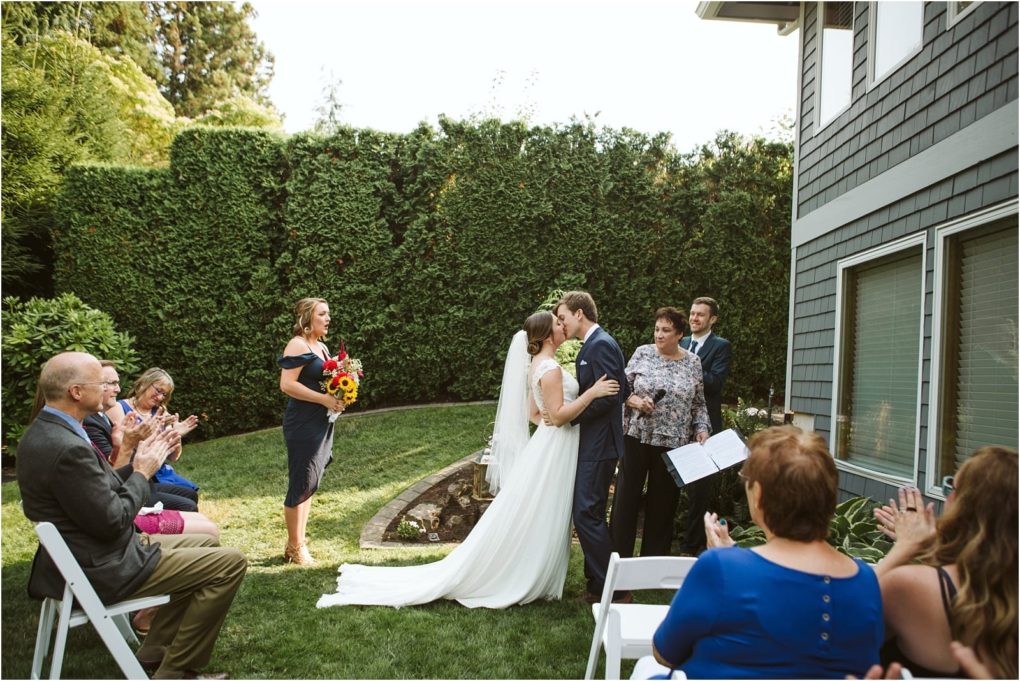 Premier snohomish & seattle wedding photographer | gsquared weddings photography a bride and groom share their first kiss at an outdoor snohomish wedding ceremony surrounded by clapping guests seated on white chairs Bridesmaid holds flowers nearby next to a tall green hedge and gray house | serving snohomish seattle                   & beyond
