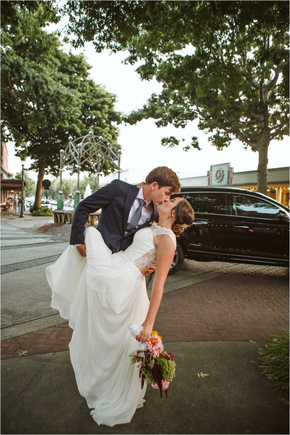 Premier snohomish & seattle wedding photographer | gsquared weddings photography a groom in a blue suit dips and kisses his bride who holds a bouquet and smiles on a sidewalk under leafy trees in snohomish with storefronts and a parked black carcapturing the joy of a seattle wedding engagement | serving snohomish seattle                   & beyond