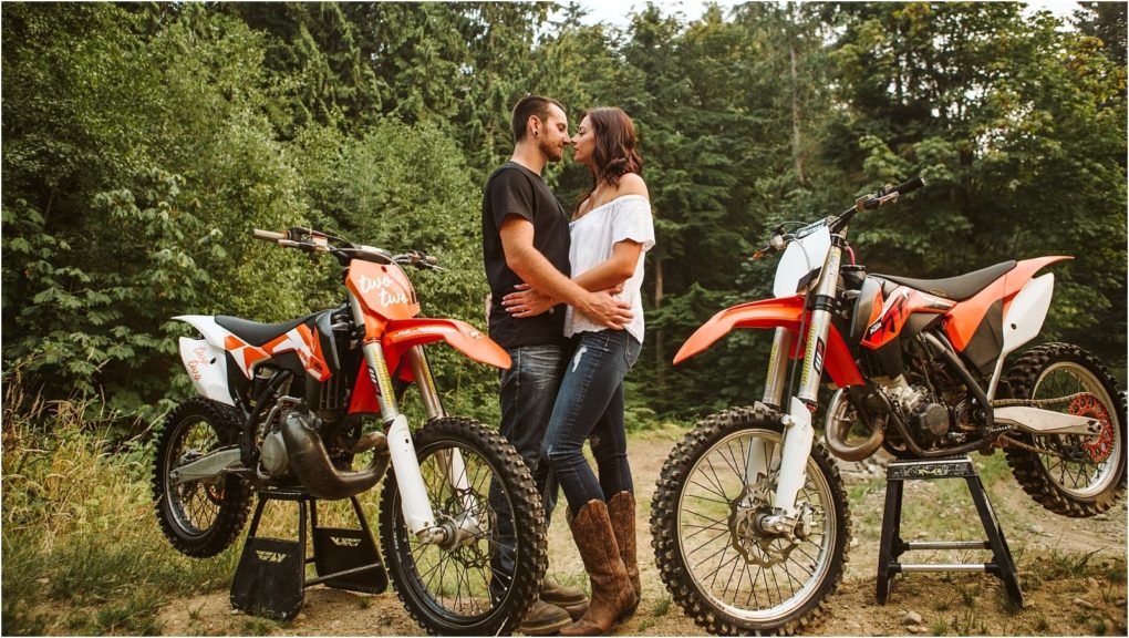 A couple stands facing each other embracing between two red dirt bikes in a forested snohomish outdoor setting The woman wears jeans and boots while the man wears a black shirt and jeans Snohomish wedding photography