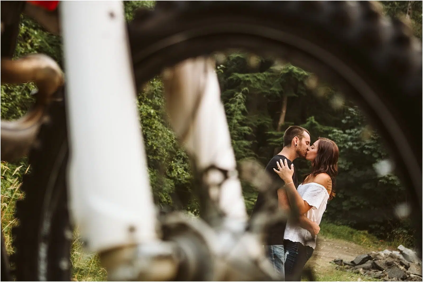 A couple kisses in a forested area near Snohomish, framed creatively through the wheel of a bicycle in the foreground. The scene is outdoors and intimate, with lush greenery surrounding them. Snohomish Wedding Photography