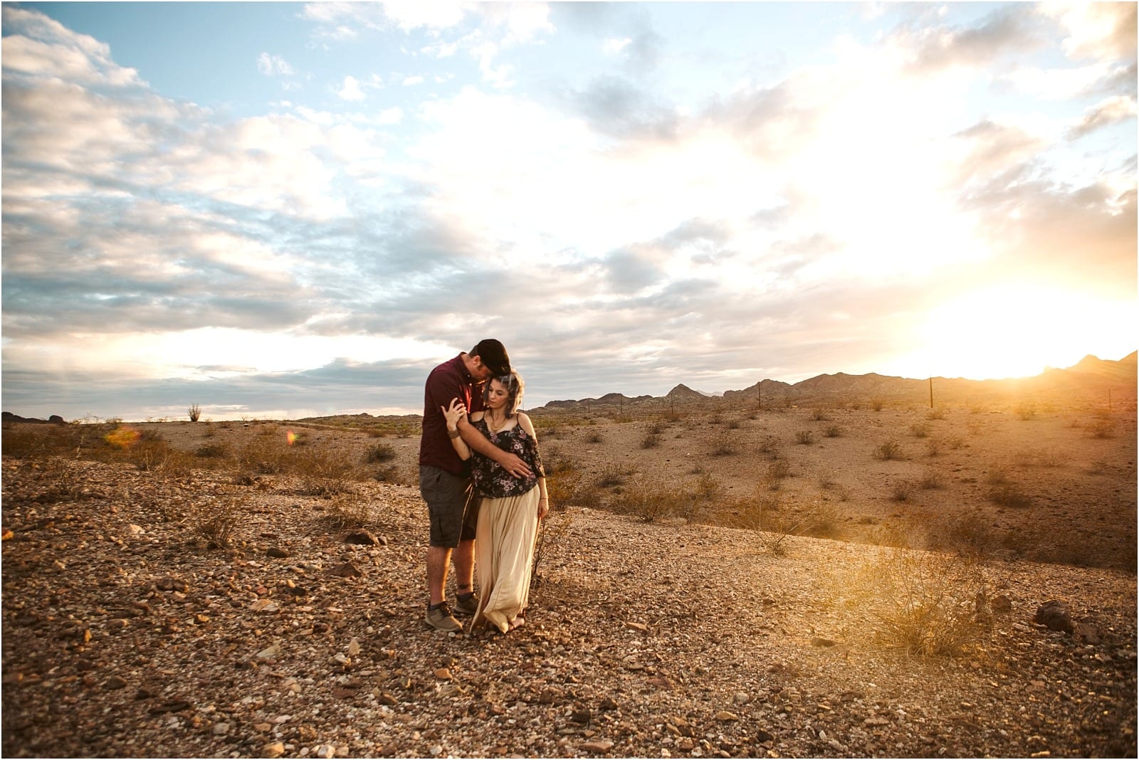 Premier Snohomish & Seattle Wedding Photographer | GSquared Weddings Photography A couple embraces in a rocky desert at sunset, with mountains and a cloudy sky in the background. Perfect for a Snohomish or Seattle wedding engagement, warm sunlight glows on the horizon as they share a romantic moment. 47.9129° N, 122.0982° W | Serving Snohomish, Seattle , Orlando & Beyond