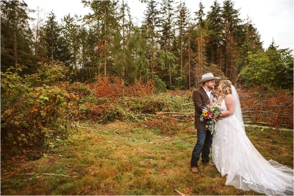 Premier snohomish & seattle wedding photographer | gsquared weddings photography a bride in a white gown and a groom in a cowboy hat embrace in a grassy clearing near snohomish surrounded by trees and greenery with the bride holding a colorful bouquetcapturing the charm of a seattle wedding engagement | serving snohomish seattle                   & beyond