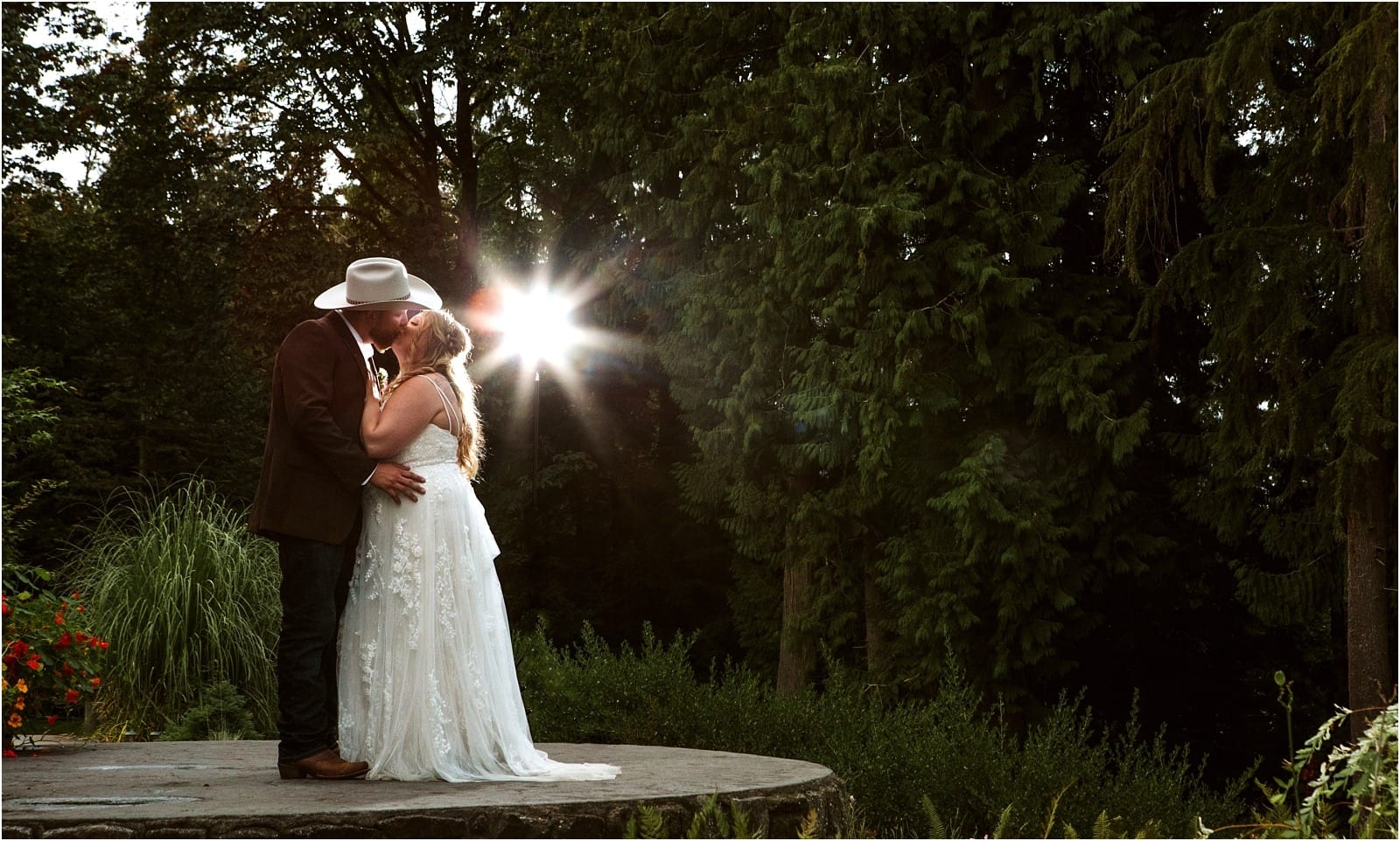 Premier Snohomish & Seattle Wedding Photographer | GSquared Weddings Photography A bride and groom share a kiss on a stone platform surrounded by lush greenery at their Snohomish wedding, sunlight streaming through the trees. The groom wears a cowboy hat; the bride is in a long white dress with loose hair.                                   | Serving Snohomish, Seattle ,                  & Beyond