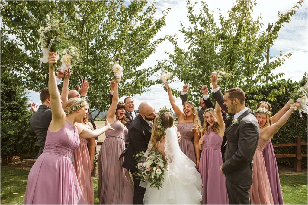 Premier snohomish & seattle wedding photographer | gsquared weddings photography a bride and groom kiss outdoors at their snohomish seattle wedding surrounded by a joyful wedding party in lavender dresses and dark suits arms raised in celebration under green trees on a sunny day | serving snohomish seattle                   & beyond