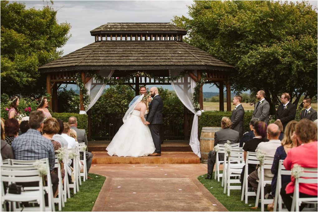 Premier snohomish & seattle wedding photographer | gsquared weddings photography a bride and groom kiss under a wooden gazebo during their snohomish outdoor wedding ceremony Guests sit on white chairs the wedding party stands nearby and lush greenery surrounds this romantic seattle engagement scene | serving snohomish seattle                   & beyond