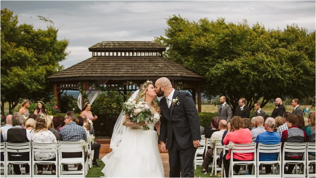 Premier snohomish & seattle wedding photographer | gsquared weddings photography a bride and groom kiss while holding hands at their snohomish outdoor wedding ceremony in front of a gazebo surrounded by seated guests and wedding party members under cloudy skies near seattle | serving snohomish seattle                   & beyond