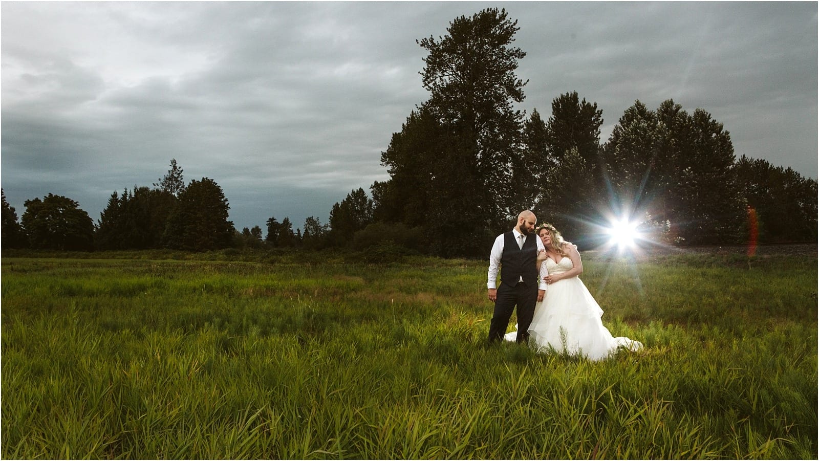 Premier Snohomish & Seattle Wedding Photographer | GSquared Weddings Photography A bride and groom stand together in a green field at dusk near Snohomish, with trees and a cloudy sky in the background. The bride leans on the groom, while a bright flash of light appears behind them during their Seattle wedding.                                   | Serving Snohomish, Seattle ,                  & Beyond