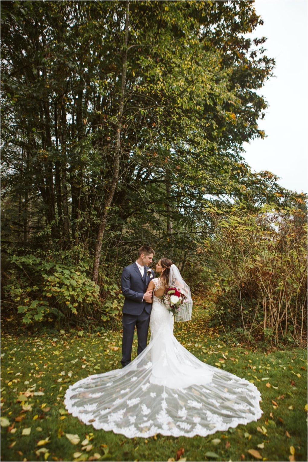 Premier snohomish & seattle wedding photographer | gsquared weddings photography a bride in a long lace wedding gown and veil stands on green grass holding a bouquet and gazing at the groom in a navy suit during their snohomish wedding They are surrounded by trees with green foliage on a cloudy day | serving snohomish seattle                   & beyond