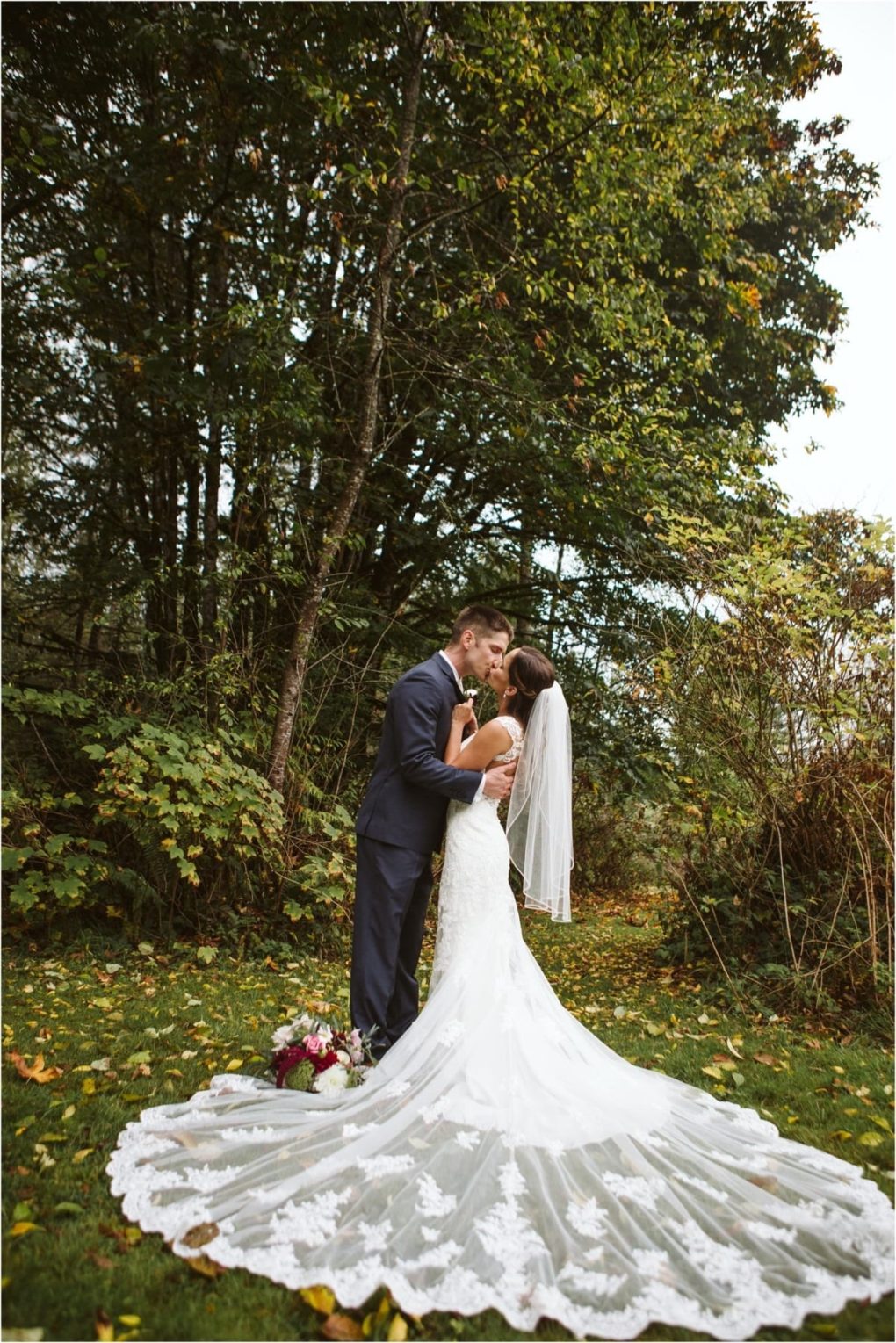 Premier snohomish & seattle wedding photographer | gsquared weddings photography a bride and groom share a kiss outdoors at their snohomish wedding surrounded by lush trees The brides white dress with a long lace train and veil shines as the groom in a dark suit stands nearby bouquet resting gently on the grass | serving snohomish seattle                   & beyond