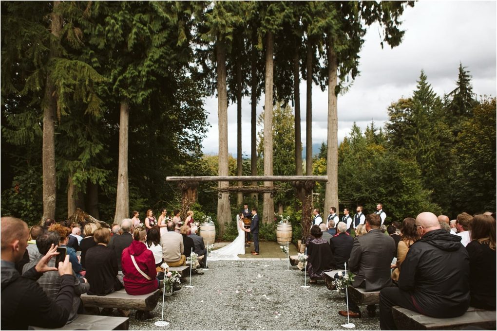 Premier snohomish & seattle wedding photographer | gsquared weddings photography an outdoor snohomish wedding ceremony is taking place in a forest clearing near seattle Guests sit on benches facing the bride and groom under a wooden arch surrounded by tall trees and lush greenery on a cloudy day | serving snohomish seattle                   & beyond