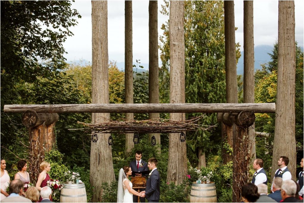 Premier snohomish & seattle wedding photographer | gsquared weddings photography a bride and groom stand facing each other holding hands during an outdoor snohomish wedding ceremony in a forest setting with tall trees and rustic wooden beams Guests and attendants stand on either side watching the couple | serving snohomish seattle                   & beyond