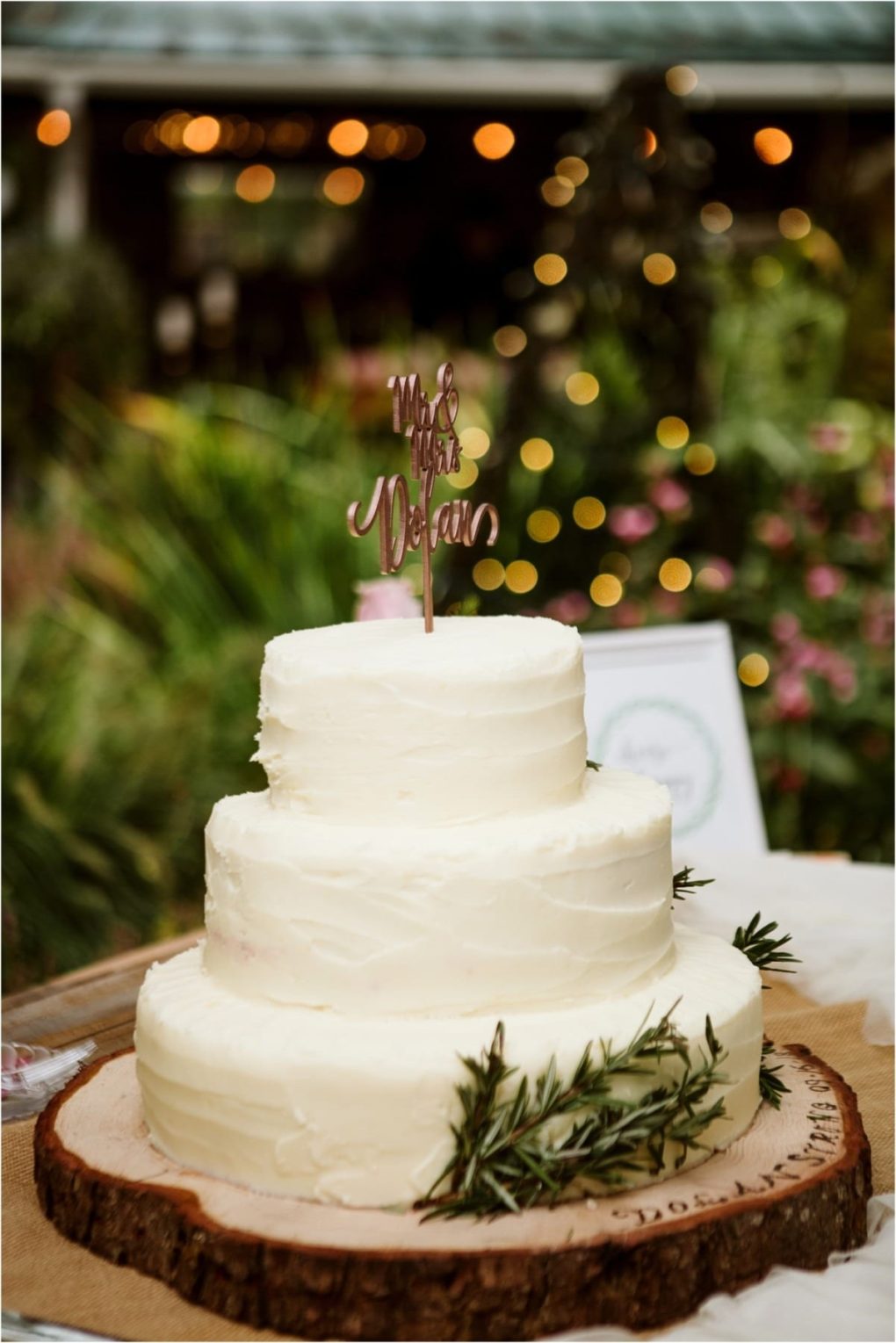 Premier snohomish & seattle wedding photographer | gsquared weddings photography a three tiered white frosted cake sits on a wooden slab decorated with greenery and a mr & mrs topperperfect for a snohomish seattle wedding or engagement set outdoors with blurred lights and lush foliage in the background | serving snohomish seattle                   & beyond
