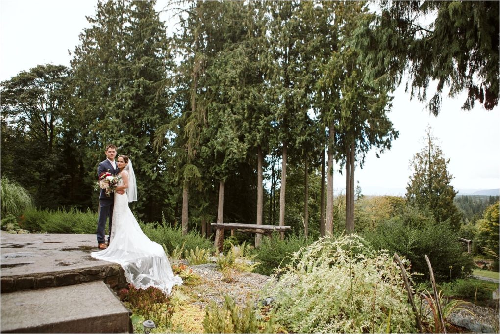 Premier snohomish & seattle wedding photographer | gsquared weddings photography a bride and groom stand together on a stone path in front of tall evergreen trees at a beautiful snohomish wedding The bride wears a long white dress and veil holding a bouquet while lush greenery surrounds the couple | serving snohomish seattle                   & beyond