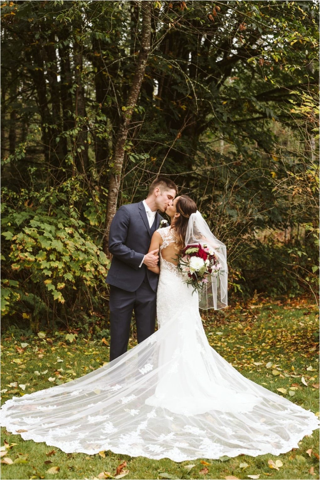 Premier snohomish & seattle wedding photographer | gsquared weddings photography a bride in a white lace gown and veil kisses her groom outdoors at their snohomish wedding She holds a bouquet of red and white flowers while her long train spreads across the grass beneath the green trees | serving snohomish seattle                   & beyond