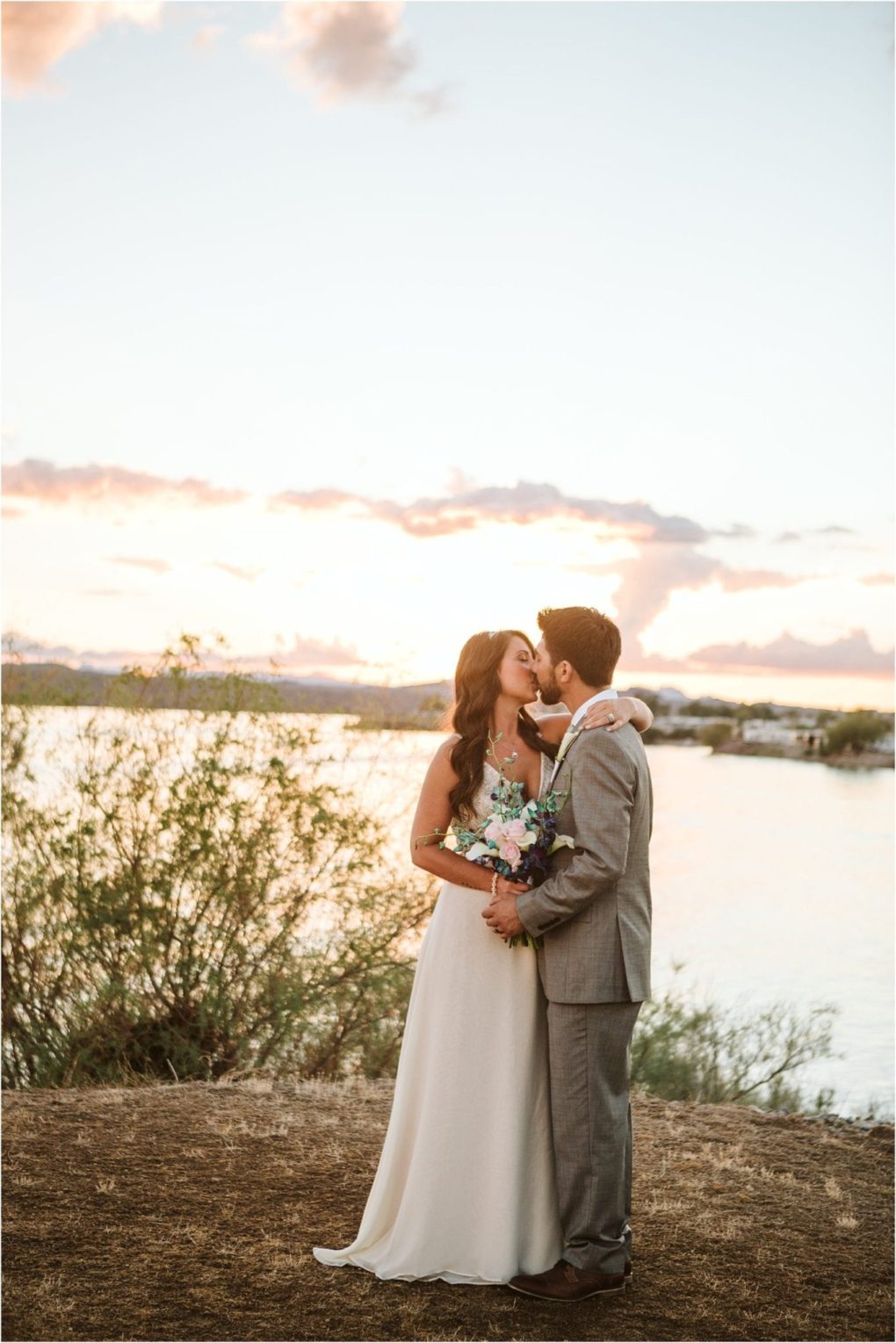 Premier snohomish & seattle wedding photographer | gsquared weddings photography a bride and groom share a kiss outdoors near a lake at sunset during their snohomish wedding The bride wears a white dress and holds a bouquet while the groom is in a gray suit Soft sunlight and clouds fill the sky in the background | serving snohomish seattle                   & beyond