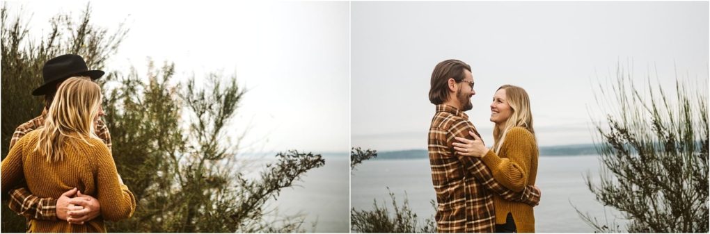 Premier snohomish & seattle wedding photographer | gsquared weddings photography a couple wearing brown plaid and mustard sweaters embraces by the water smiling at each other with greenery in the background under an overcast sky during their snohomish seattle engagement | serving snohomish seattle                   & beyond
