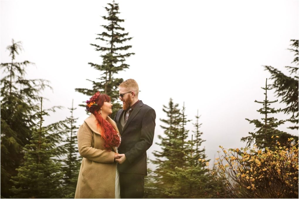 Premier snohomish & seattle wedding photographer | gsquared weddings photography a couple stands outdoors among evergreen trees in snohomish gazing at each other and smiling The woman has long red hair with a floral crown and tan coat while the man wears glasses and a dark suit under the overcast skya dreamy seattle engagement | serving snohomish seattle                   & beyond
