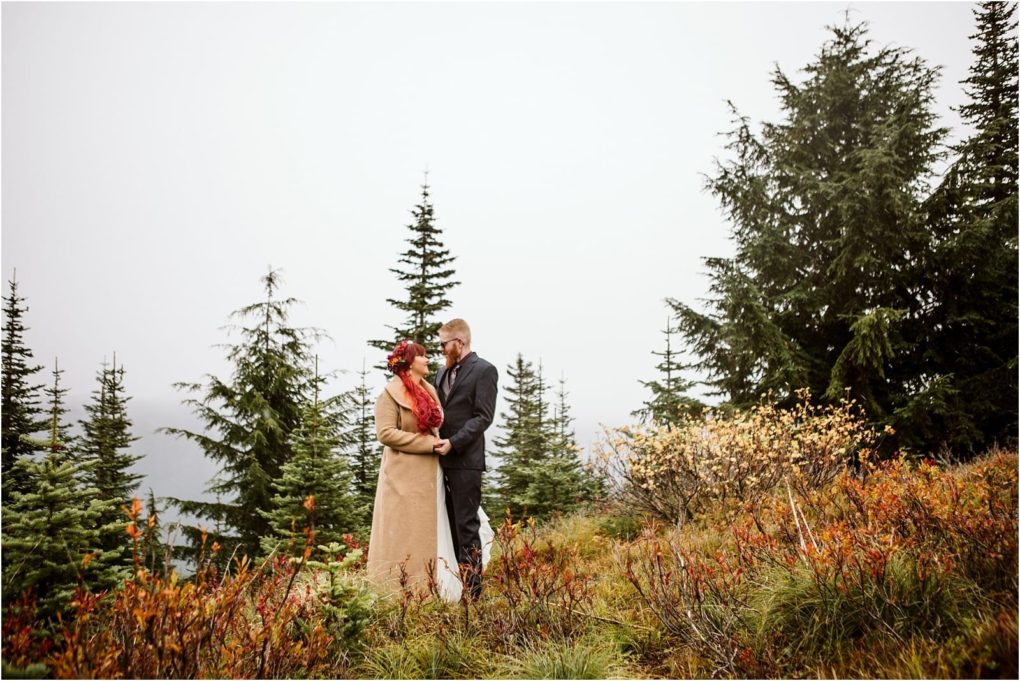 Premier snohomish & seattle wedding photographer | gsquared weddings photography a couple stands closely together on a grassy colorful snohomish hillside surrounded by evergreens The woman in a tan coat and red scarf holds a bouquet the man in a dark suit The overcast sky sets a romantic seattle engagement scene | serving snohomish seattle                   & beyond