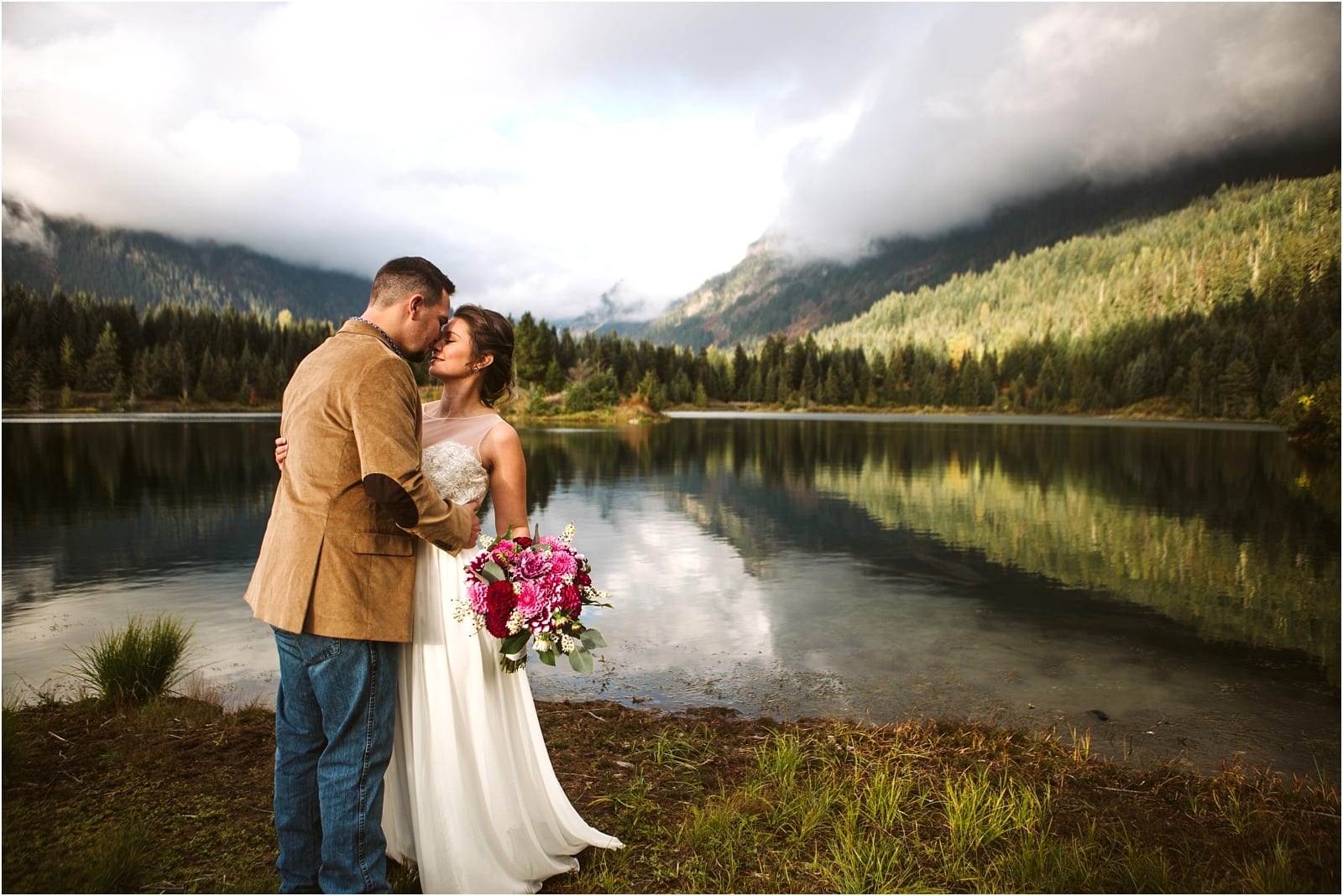 Premier Snohomish & Seattle Wedding Photographer | GSquared Weddings Photography A bride and groom stand close together by a calm lake near Snohomish, surrounded by trees and mountains, with the bride holding a bouquet of pink flowers. The partly cloudy sky creates a serene, romantic Seattle wedding scene.                                   | Serving Snohomish, Seattle ,                  & Beyond