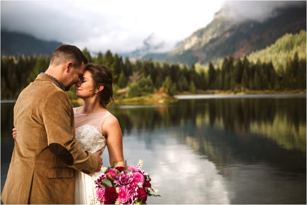 Premier snohomish & seattle wedding photographer | gsquared weddings photography a snohomish bride and groom embrace by a calm lake with pine trees and mountains in the background The bride holds a bouquet of pink flowers and both appear peaceful and in love on their seattle wedding day | serving snohomish seattle                   & beyond