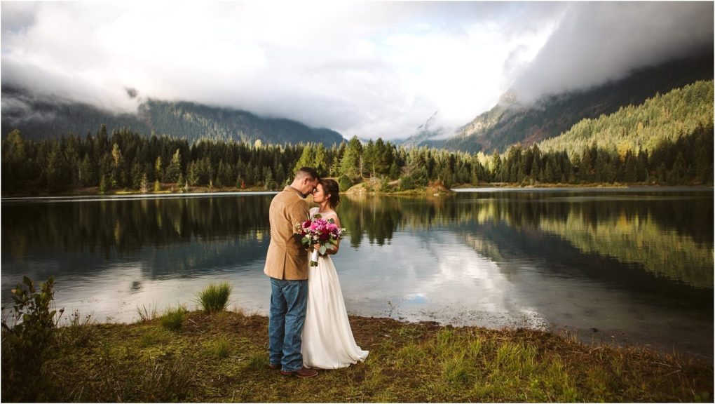 Premier snohomish & seattle wedding photographer | gsquared weddings photography a bride and groom stand embracing by a calm lake near snohomish surrounded by pine trees and mountains under a cloudy sky The bride holds a bouquet their reflection shimmeringa perfect seattle wedding engagement moment | serving snohomish seattle                   & beyond