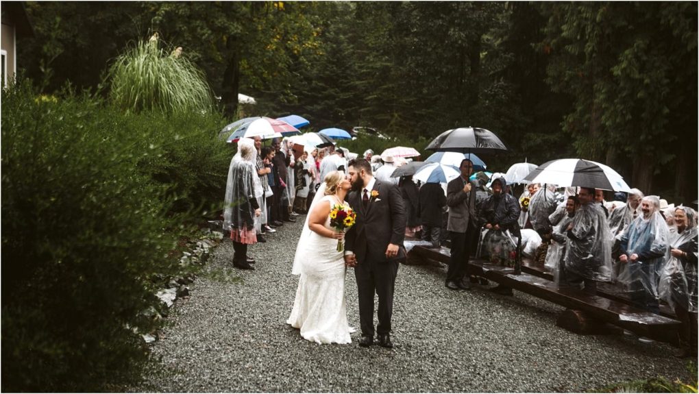 Premier snohomish & seattle wedding photographer | gsquared weddings photography a bride and groom share a kiss outdoors on a rainy snohomish wedding day surrounded by guests holding umbrellas and wearing rain ponchos The lush green setting near seattle features benches filled with joyful friends and family | serving snohomish seattle                   & beyond