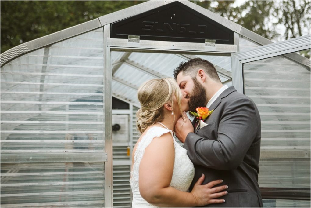 Premier snohomish & seattle wedding photographer | gsquared weddings photography a bride and groom share a kiss outside a small greenhouse at their snohomish seattle wedding with the bride in a lace dress and the groom in a suit with an orange boutonniere The greenhouse door is open behind them | serving snohomish seattle                   & beyond
