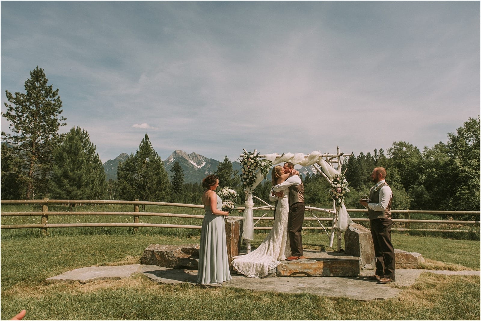 Premier Snohomish & Seattle Wedding Photographer | GSquared Weddings Photography A bride and groom share a kiss at their Snohomish outdoor wedding ceremony, standing under a decorated arch with mountains and pine trees in the background. Two attendants, one in a silver dress and one in a vest, stand nearby.                                   | Serving Snohomish, Seattle ,                  & Beyond