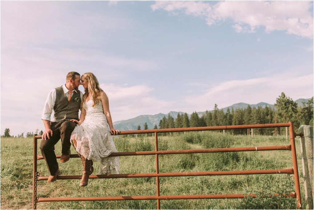 Premier snohomish & seattle wedding photographer | gsquared weddings photography a bride and groom sit on a rusty metal gate in a grassy field near snohomish sharing a kiss The bride wears a white dress and boots the groom wears a vest and tie Mountains and trees are visible under the blue seattle sky | serving snohomish seattle                   & beyond