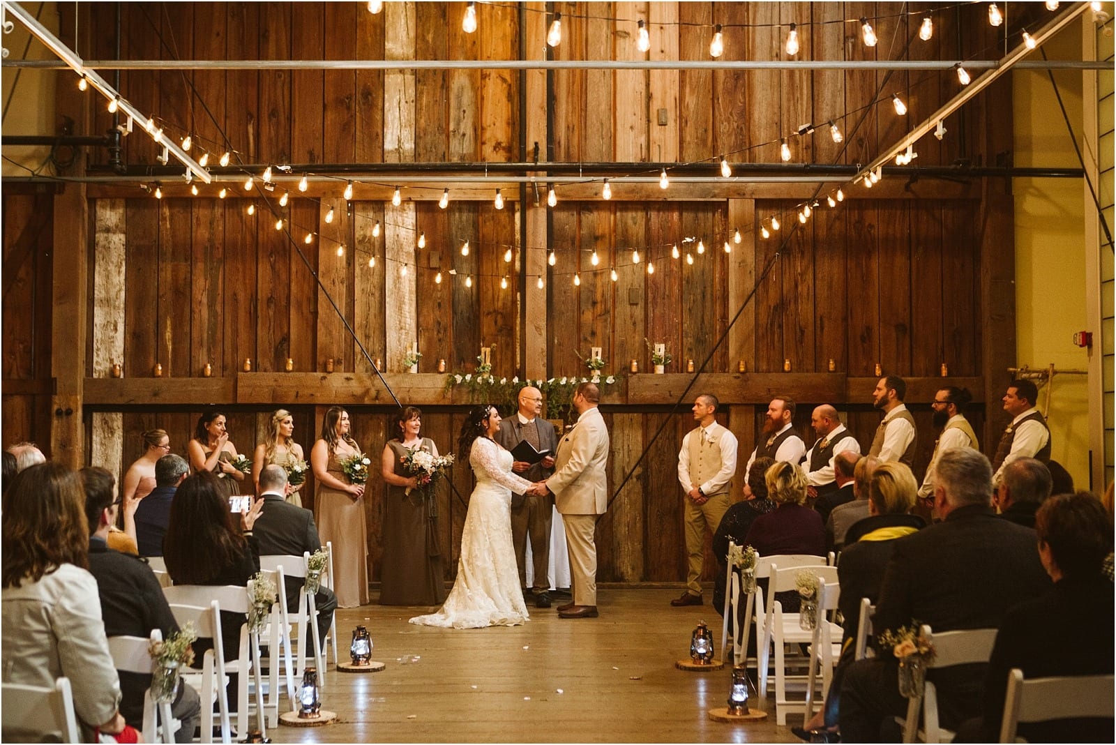 Premier Snohomish & Seattle Wedding Photographer | GSquared Weddings Photography A bride and groom stand holding hands at the altar during an indoor rustic Snohomish wedding ceremony, surrounded by their wedding party and guests, beneath string lights and in front of a wooden backdrop.                                   | Serving Snohomish, Seattle ,                  & Beyond