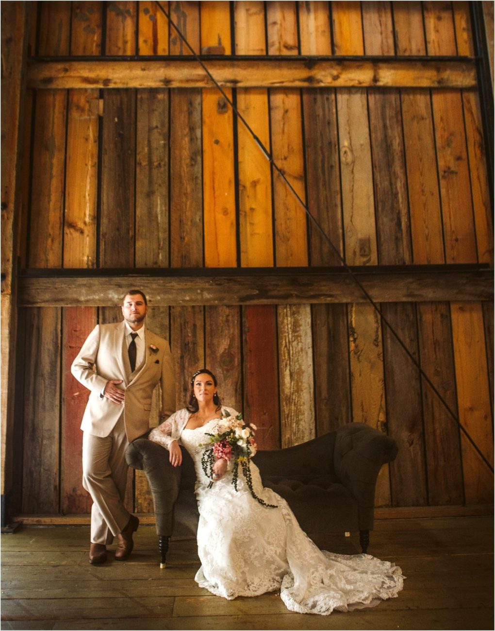 Premier snohomish & seattle wedding photographer | gsquared weddings photography a bride in a white lace gown sits on a dark sofa holding a bouquet while her groom in a beige suit stands beside her They pose against a large rustic wooden wallcapturing the warmth of a snohomish seattle wedding engagement | serving snohomish seattle                   & beyond