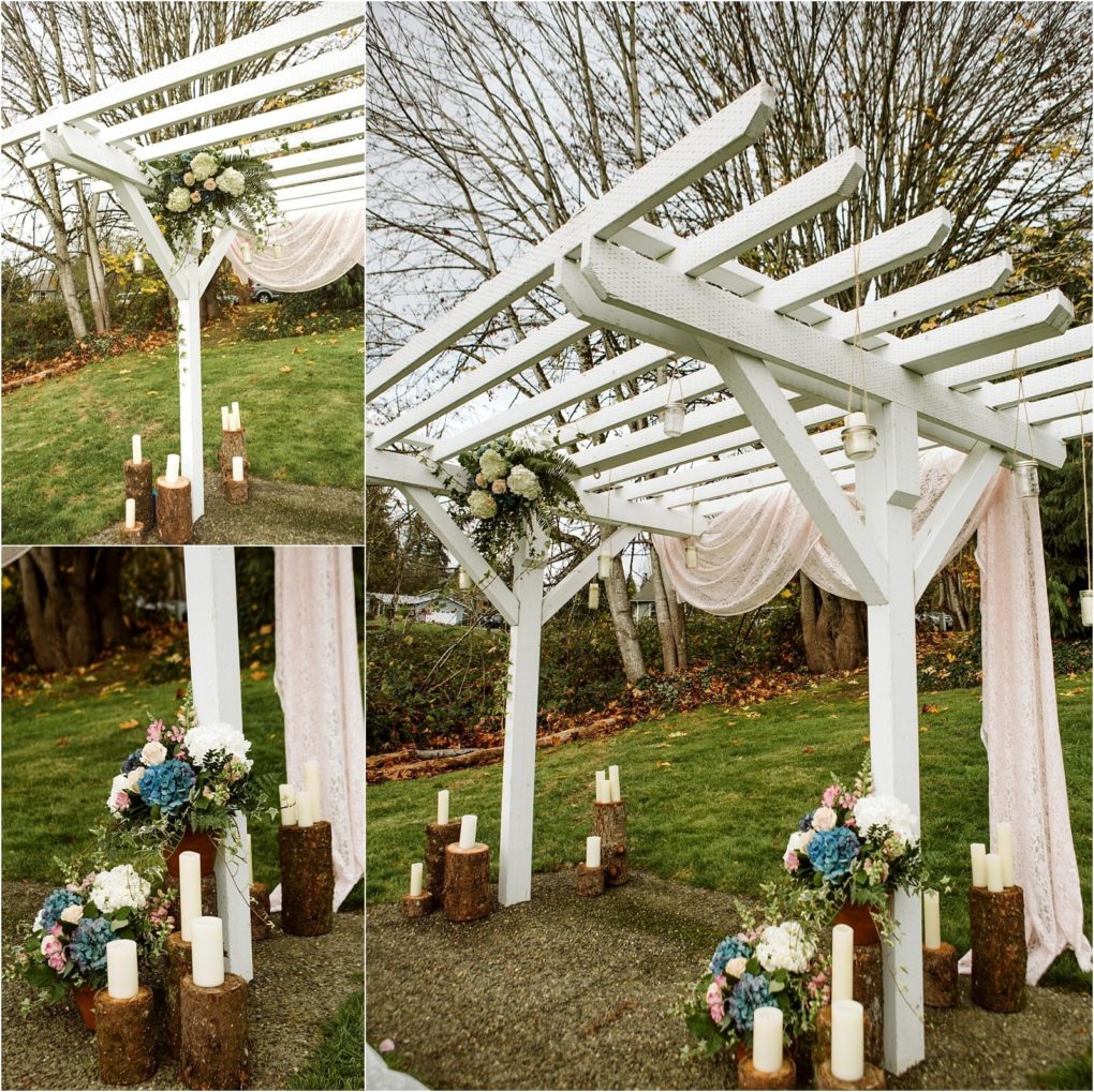 Premier snohomish & seattle wedding photographer | gsquared weddings photography a white wooden wedding arch decorated with pastel flowers greenery hanging candles and draped pink fabric stands outdoors on grass for a romantic snohomish engagement Logs and pillar candles rest at the base trees and overcast sky behind | serving snohomish seattle                   & beyond
