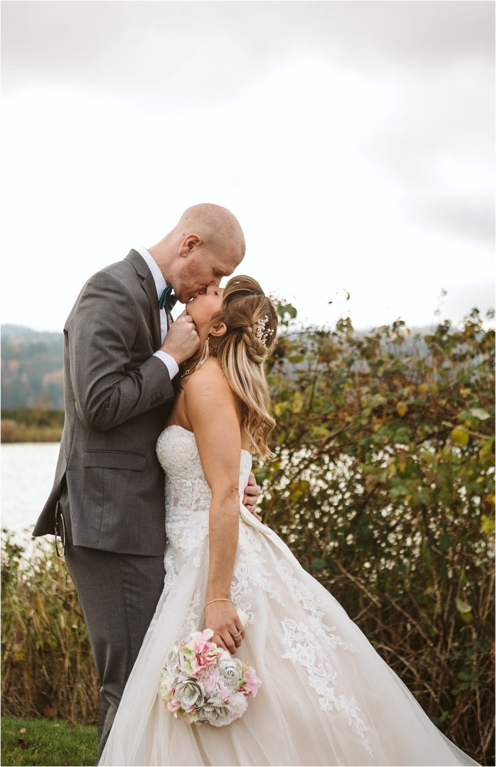 Premier snohomish & seattle wedding photographer | gsquared weddings photography a groom in a gray suit gently kisses the forehead of his bride in a white gown holding a bouquet standing outdoors with greenery and watera romantic snohomish seattle wedding engagement moment | serving snohomish seattle                   & beyond