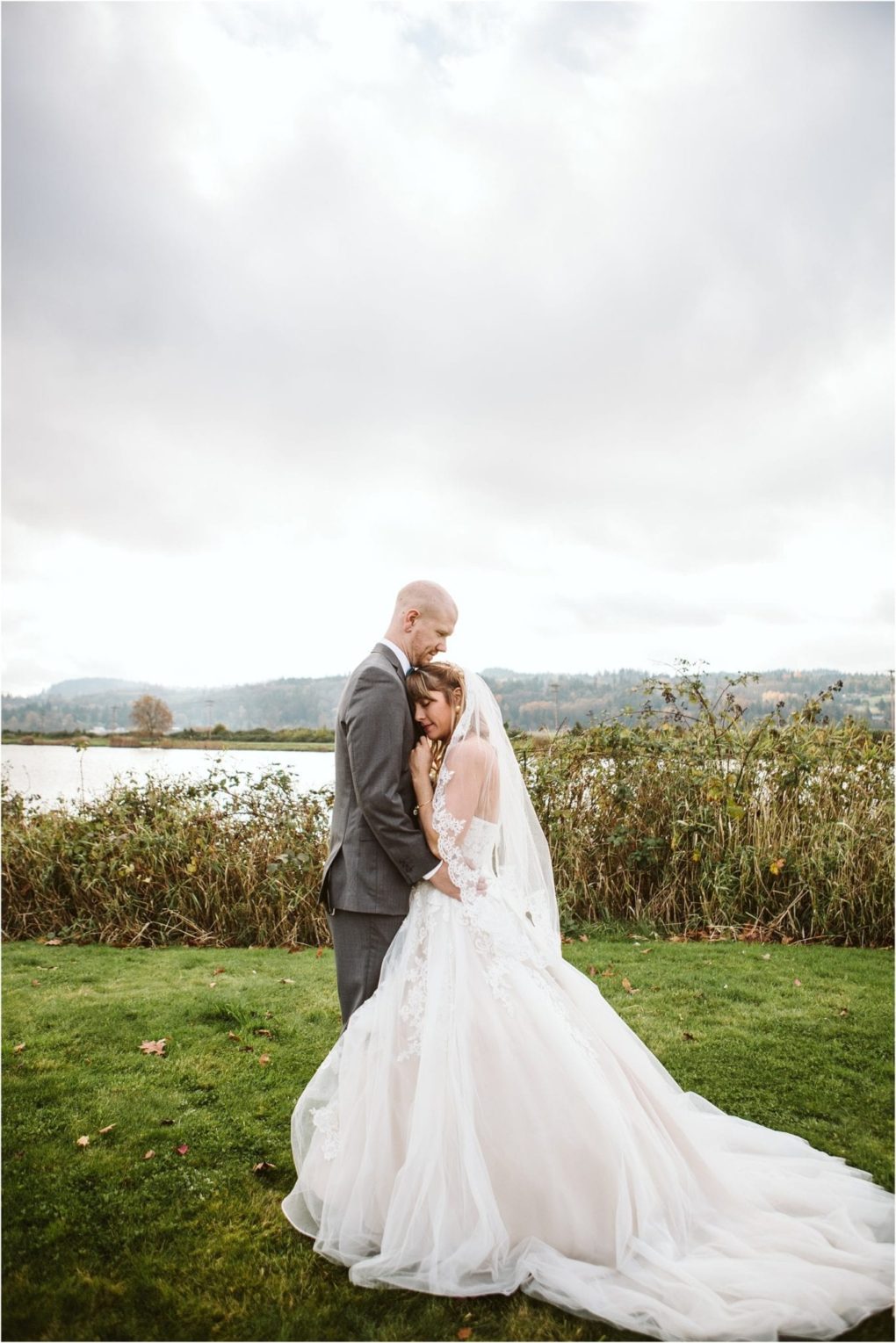 Premier snohomish & seattle wedding photographer | gsquared weddings photography a bride in a flowing white gown and veil embraces a groom in a gray suit on a grassy field with a lake and cloudy sky in the background capturing the romance of a snohomish seattle wedding engagement | serving snohomish seattle                   & beyond