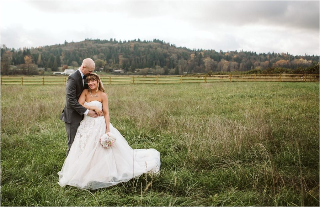 Premier snohomish & seattle wedding photographer | gsquared weddings photography a bride and groom stand together in a grassy field near snohomish with hills and trees in the background The groom hugs the bride from behind as she smiles holding a bouquet and wearing a strapless white gown under the cloudy sky | serving snohomish seattle                   & beyond