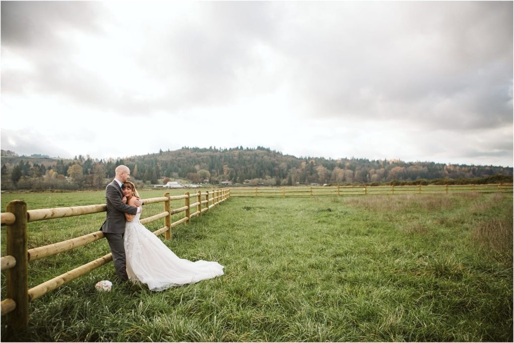 Premier snohomish & seattle wedding photographer | gsquared weddings photography a bride and groom embrace beside a wooden fence in a grassy field near snohomish with rolling hills and trees under a cloudy sky The brides bouquet rests on the grasscapturing pure seattle wedding romance | serving snohomish seattle                   & beyond