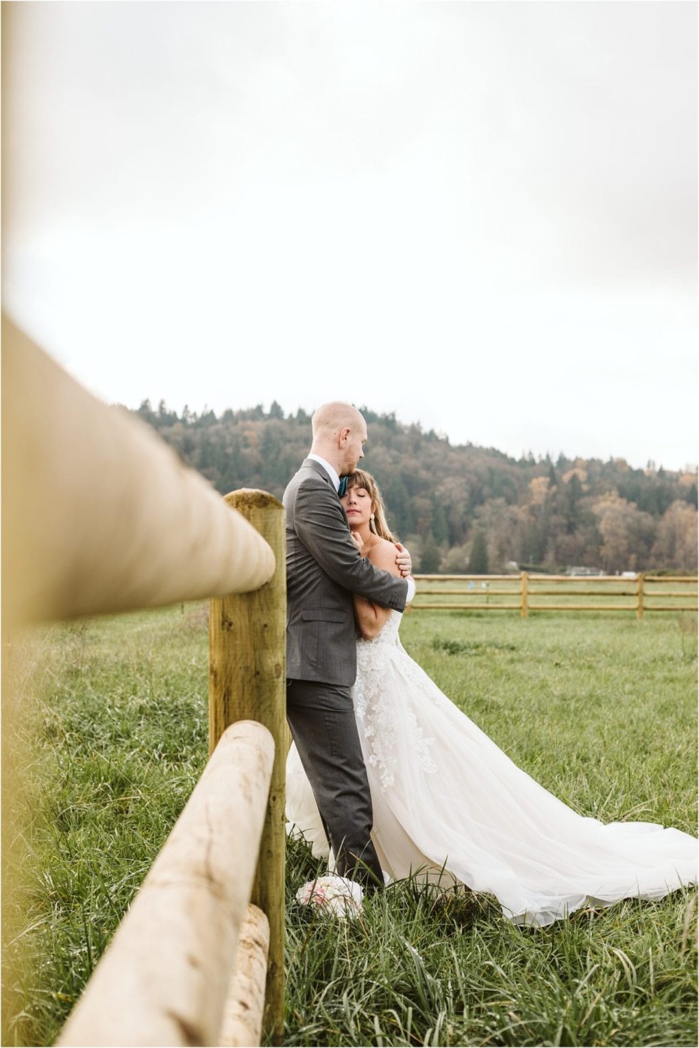 Premier snohomish & seattle wedding photographer | gsquared weddings photography a bride in a white gown and a groom in a suit embrace beside a wooden fence in a grassy field with forested hills behind them under a cloudy skycapturing the magic of a snohomish seattle wedding engagement | serving snohomish seattle                   & beyond