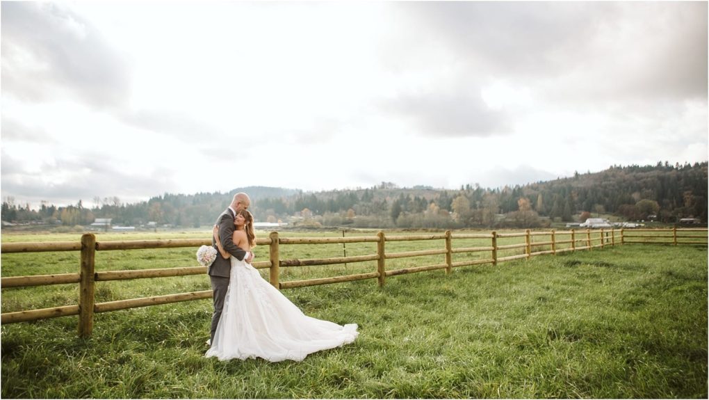 Premier snohomish & seattle wedding photographer | gsquared weddings photography a snohomish bride and groom embrace in a grassy field next to a wooden fence with trees and hills in the background under a cloudy sky The bride wears a white gown and holds a bouquet perfect for a seattle wedding engagement | serving snohomish seattle                   & beyond