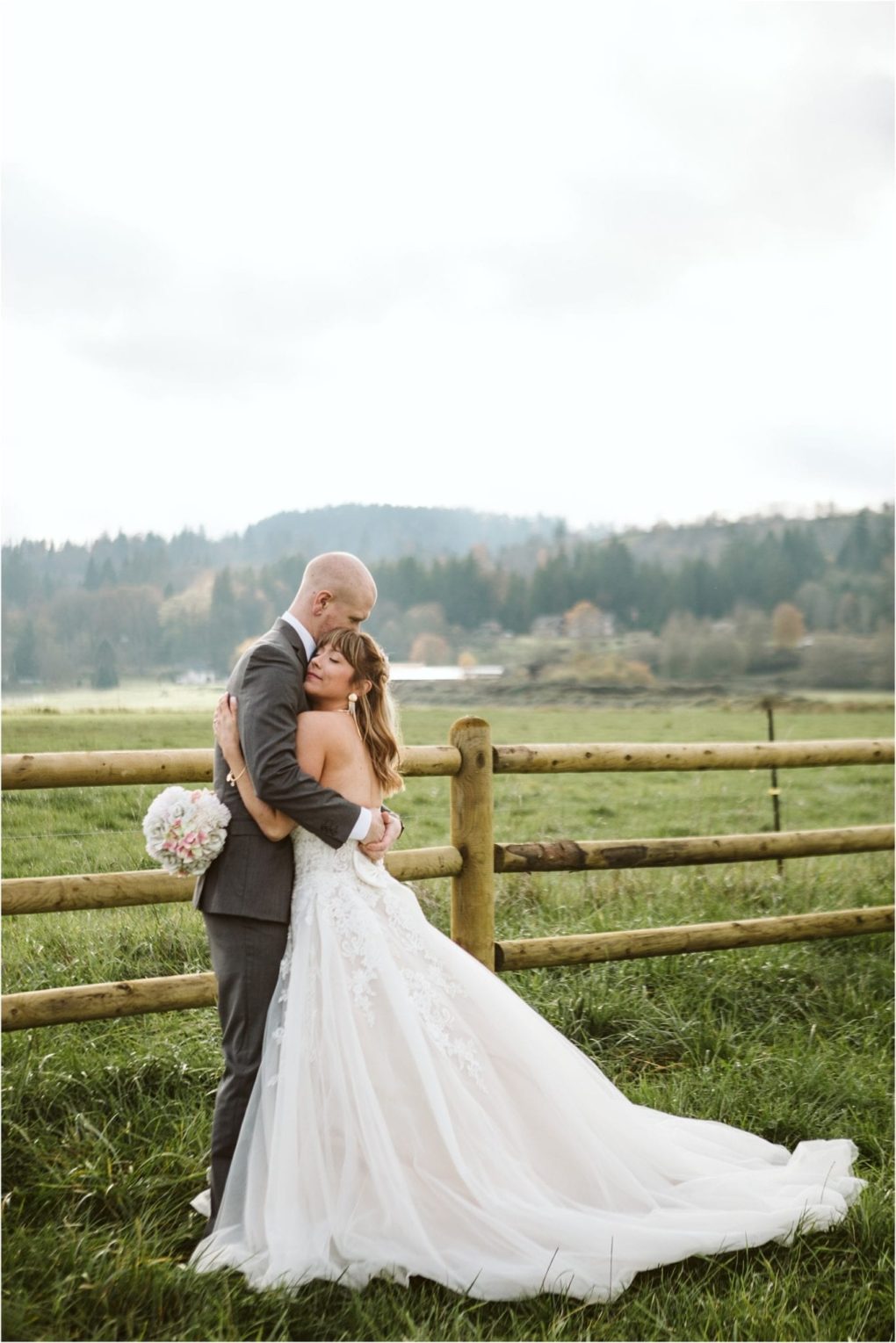 Premier snohomish & seattle wedding photographer | gsquared weddings photography a bride in a white gown and groom in a suit embrace lovingly by a wooden fence with snohomishs green fields and forested hills behind themcapturing the charm of a seattle wedding engagement The bride holds a bouquet of white and pink flowers | serving snohomish seattle                   & beyond