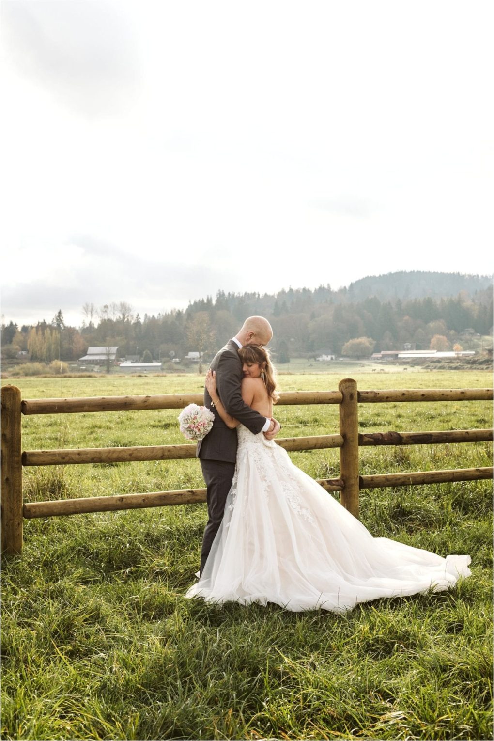 Premier snohomish & seattle wedding photographer | gsquared weddings photography a bride and groom embrace in front of a wooden fence in a grassy field with forested hills and a cloudy sky behind themcapturing the romance of a snohomish seattle wedding The bride holds a bouquet and wears a long white dress | serving snohomish seattle                   & beyond
